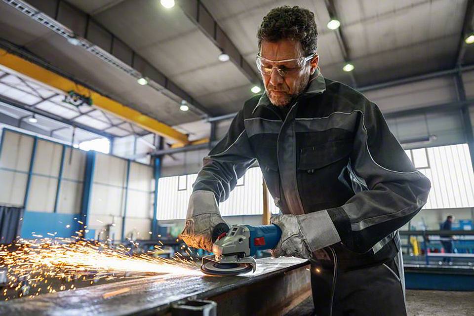 Ein Arbeiter in einer Fabrik schleift Metall mit einem Winkelschleifer, Funken fliegen. Er trägt Schutzkleidung und Schutzbrille.