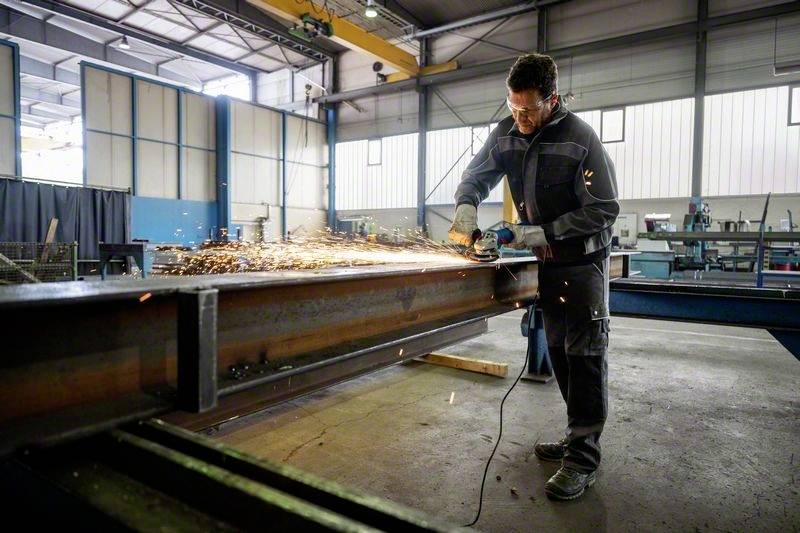 Ein Arbeiter verwendet einen Winkelschleifer, um Metall in einer Fabrikhalle zu bearbeiten. Funken fliegen in alle Richtungen.