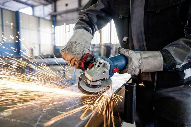 Eine Person in Schutzkleidung schleift mit einem Winkelschleifer Metall, Funken fliegen. Die Szene findet in einer Werkstatt statt.