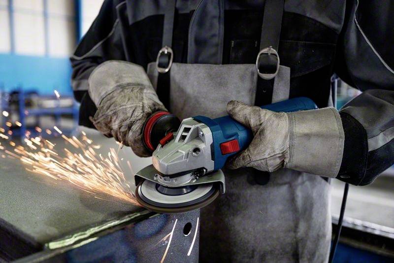 Ein Arbeiter schleift Metall mit einem Winkelschleifer. Funken sprühen vom Werkstück. Der Arbeiter trägt Schutzkleidung und Handschuhe.
