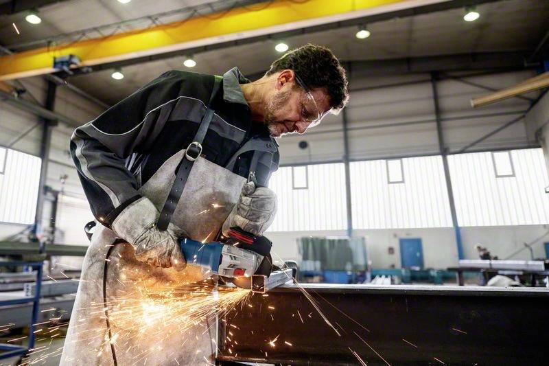 Ein Arbeiter in Schutzkleidung schleift Metall mit einem Winkelschleifer. Funken fliegen, während er in einer Fabrikumgebung arbeitet.