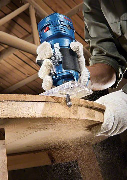 Eine Person bearbeitet mit einer Fräse die Kante eines Holztisches. Sägespäne fliegen, während die Person weiße Handschuhe trägt.