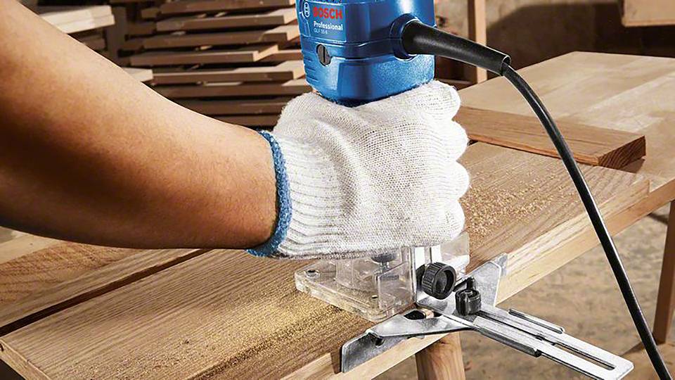 Person mit weißen Arbeitshandschuhen führt eine blaue Fräsmaschine über ein Holzstück. Holzspäne sind sichtbar.