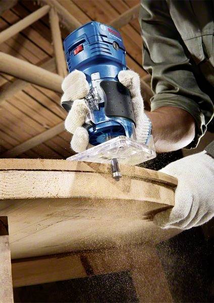 Eine Person fräst eine Holzkante mit einem blauen Fräsgerät. Holzspäne fliegen, während die Kante bearbeitet wird.