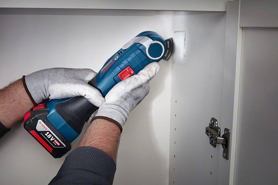 Eine Person arbeitet mit einem blauen, akkubetriebenen Multitool in einem weißen Schrank, trägt graue Handschuhe.