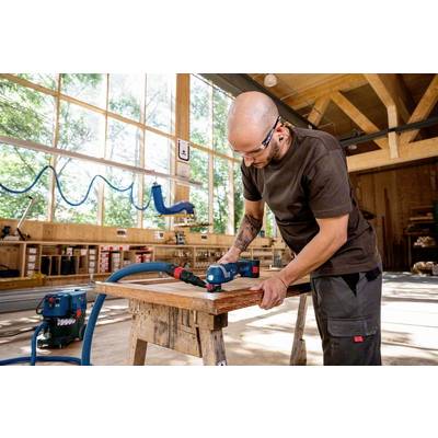 Eine Person in einer Werkstatt verwendet ein elektrisches Werkzeug, um Holz zu bearbeiten. Große Fenster im Hintergrund lassen Tageslicht herein.