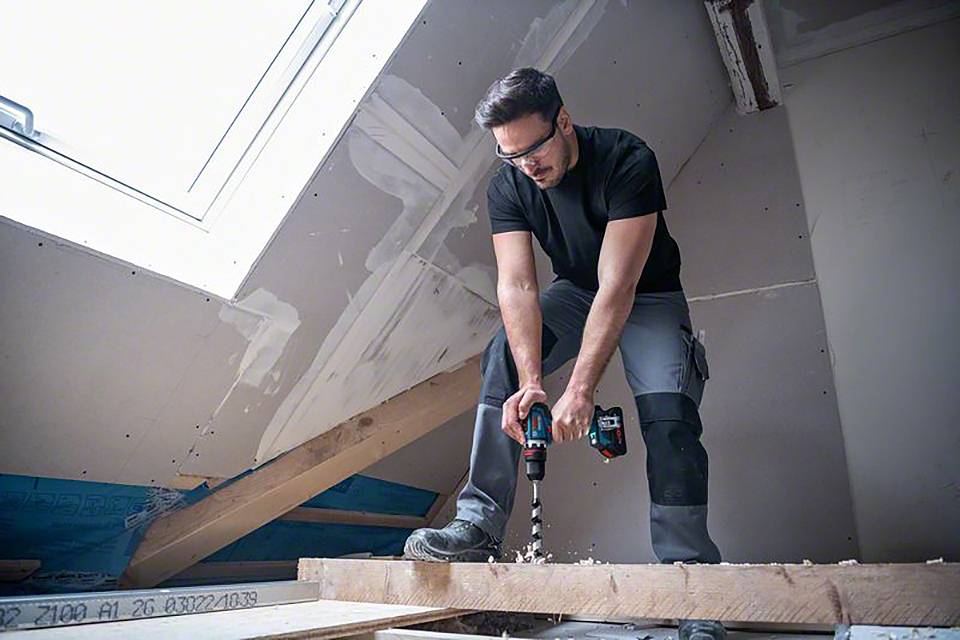 Ein Mann arbeitet auf einer Baustelle im Dachgeschoss und bohrt mit einem Akku-Bohrschrauber ein Loch in einen Holzbalken.