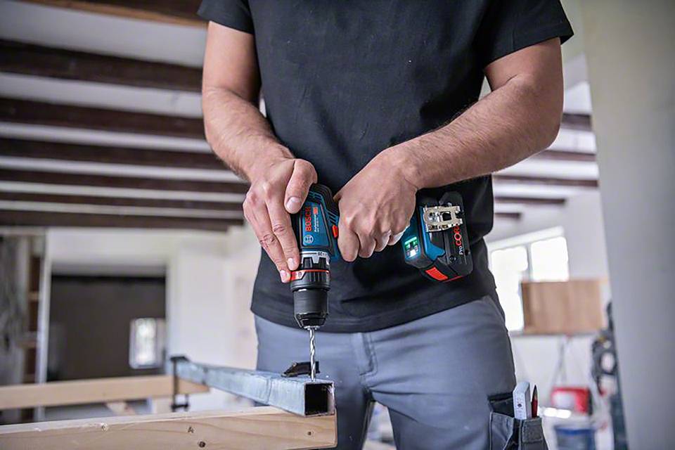 Eine Person bohrt mit einer elektrischen Bohrmaschine durch ein Metallstück in einer Werkstattumgebung.