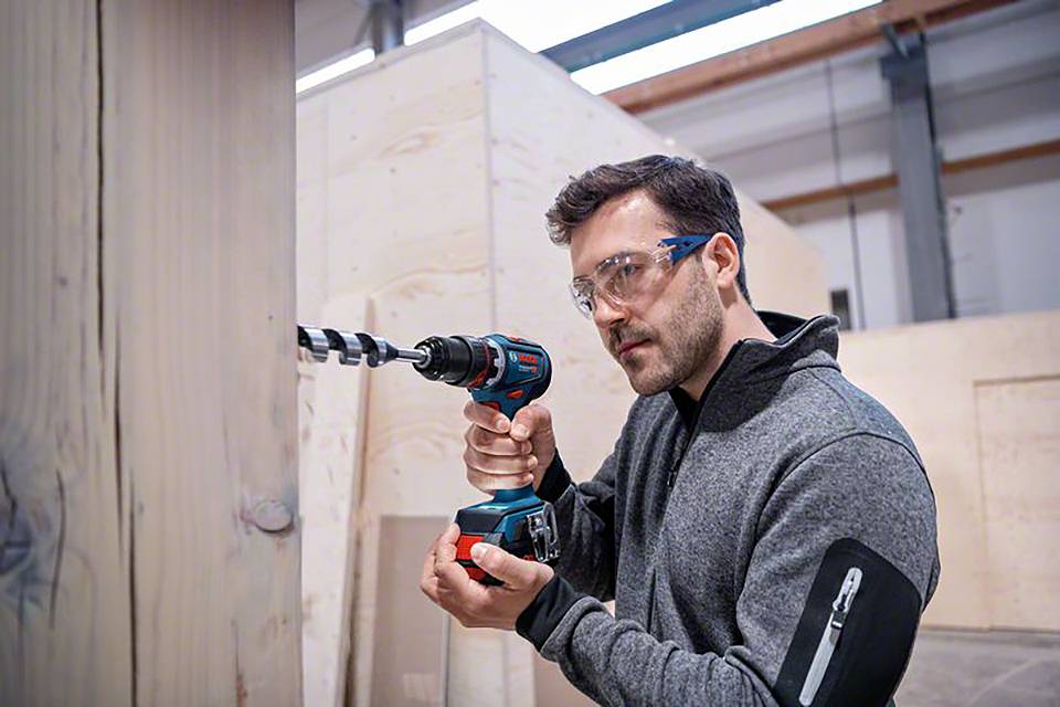 Mann in Schutzbrille bohrt mit einem Akku-Bohrschrauber in eine Holzplatte, in einer Werkstattumgebung.