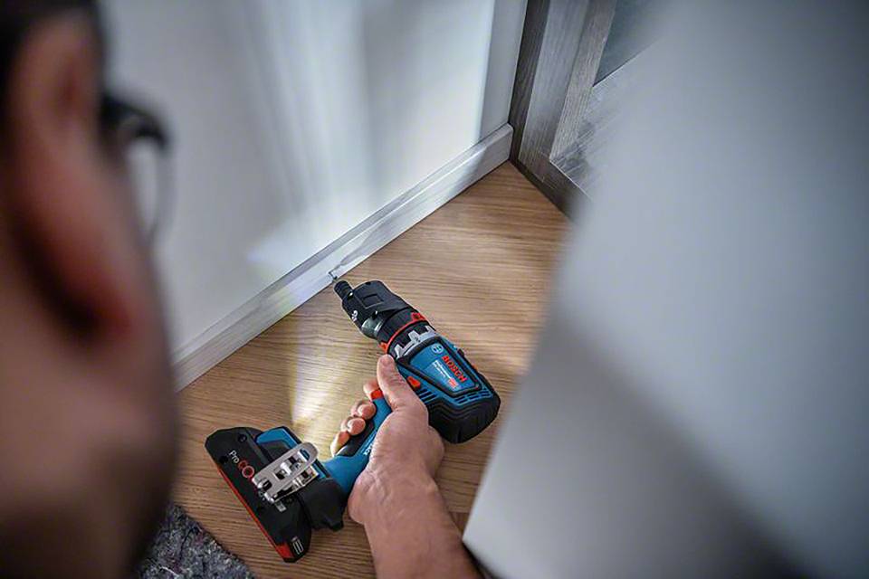 Eine Person verwendet einen Akkubohrer, um in einem Holzboden zu bohren. Die Szene ist aus der Perspektive der Person aufgenommen.