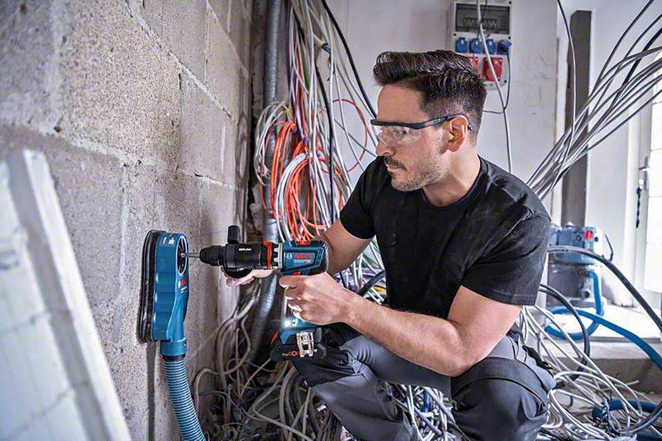 Ein Mann bohrt mit einer Bohrmaschine in eine Betonwand. Er trägt eine Schutzbrille. Im Hintergrund sind viele Kabel zu sehen.
