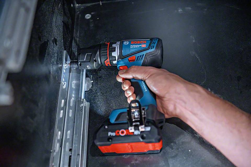 Eine Hand hält einen blauen Akkuschrauber und befestigt ein Metallscharnier an einer Wand.