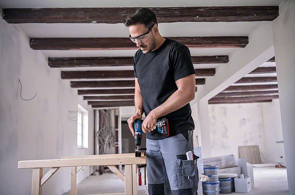 Handwerker im schwarzen T-Shirt und grauer Hose bohrt mit einer Bohrmaschine in ein Holzbrett in einem renovierungsbedürftigen Raum.