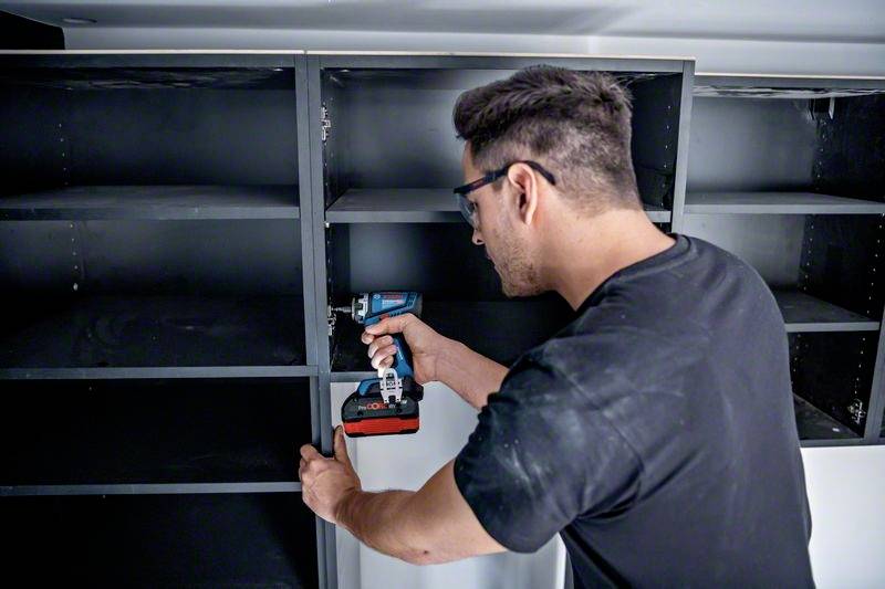 Ein Mann in schwarzem T-Shirt schraubt mit einem Akkuschrauber an einem unmontierten Schrank in einem Raum.