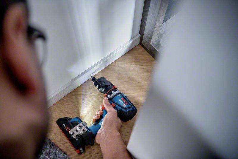 Eine Person benutzt ein Akkuschrauber, um eine Schraube in ein Holzstück in der Ecke eines Raums hineinzudrehen.