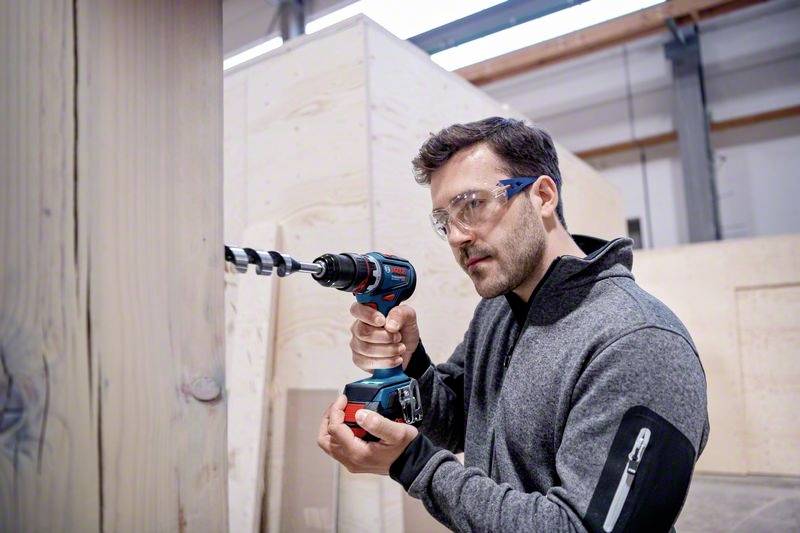 Ein Mann bohrt mit einer elektrischen Bohrmaschine ein Loch in eine Holzplatte. Er trägt Schutzbrille und Arbeitskleidung in einer Werkstatt.