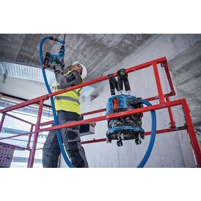 Arbeiter auf Baugerüst verwendet Bohrmaschine, trägt Schutzkleidung und -helm. Staubsauger an Gerüst befestigt, moderner Bauprozess.