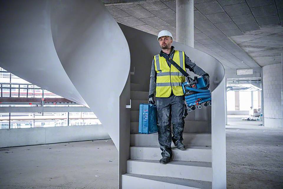 Ein Bauarbeiter mit Schutzhelm und Warnweste geht eine Treppe in einem unfertigen Gebäude hinunter, Werkzeug in der Hand.