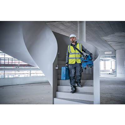 Ein Bauarbeiter mit Schutzhelm und Warnweste geht eine Treppe in einem unfertigen Gebäude hinunter, Werkzeug in der Hand.