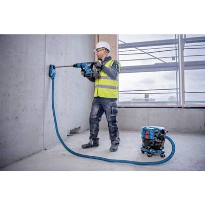Eine Person in Arbeitskleidung bohrt in eine Betonwand auf einer Baustelle. Ein Staubsauger ist an die Bohrmaschine angeschlossen.