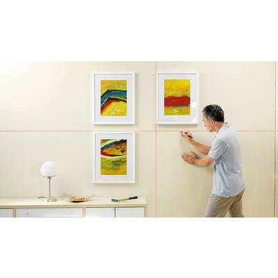 Eine Person hängt drei abstrakte Gemälde an eine beige Wand. Auf dem Schrank stehen eine Lampe und Schalen.