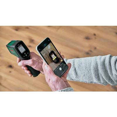 Eine Person hält ein Infrarot-Thermometer und ein Smartphone, das den Bildschirm des Thermometers fotografiert. Hintergrund ist Holzboden.