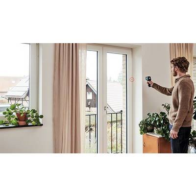 Ein Mann misst mit einem Gerät die Isolierung an einem großen Fenster. Pflanzen stehen auf der Fensterbank und einem Schrank.