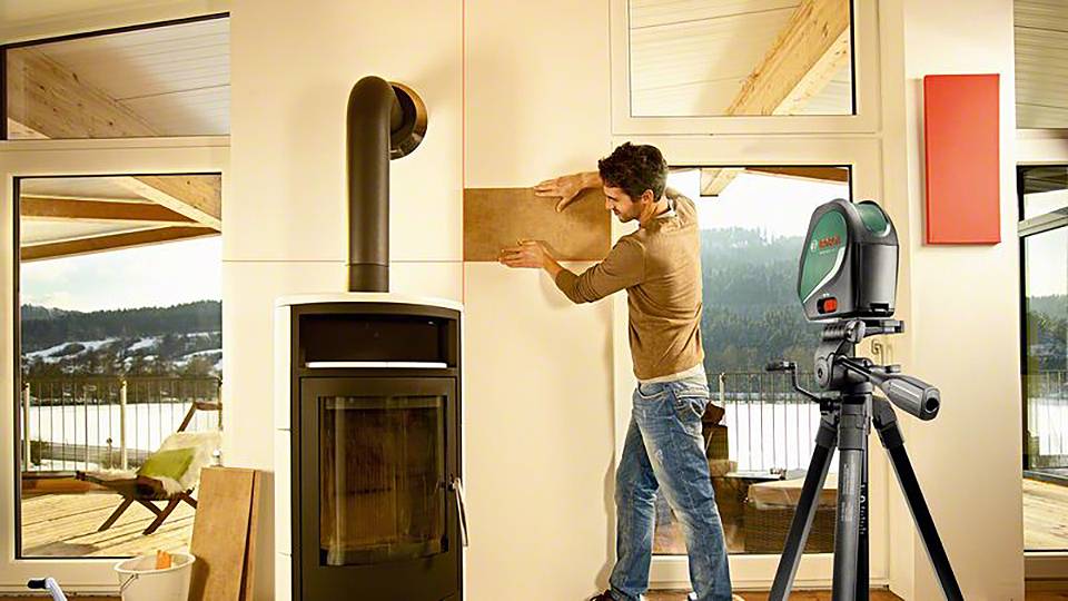 Eine Person richtet eine Holzplatte an einer Wand mit einem Laser-Entfernungsmesser aus. Im Hintergrund sind große Fenster mit Bergblick.