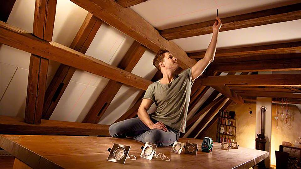 Mann sitzt auf einem Tisch in einem Dachgeschoss und weist mit einem Gerät zur Decke. Um ihn herum liegen weitere technische Geräte.