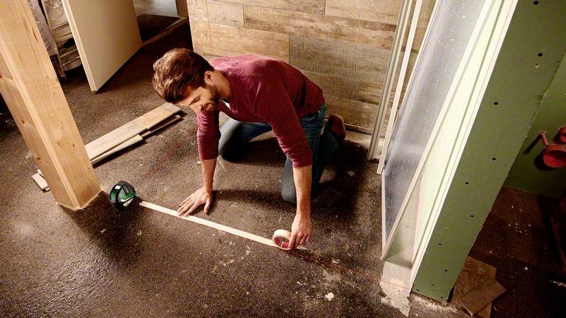 Mann auf Baustelle kniet und misst mit Maßband den Boden. Holzlatten und Werkzeuge liegen um ihn herum. Hintergrund aus Holzverkleidung.