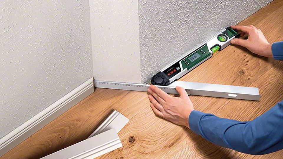 Eine Person misst mit einer Wasserwaage und einem Winkelmaß den Abstand zwischen Wand und Boden, um eine genaue Verlegung sicherzustellen.