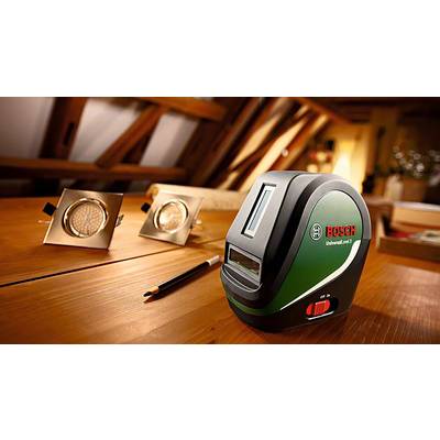 Ein grüner Bosch-Laser-Entfernungsmesser auf einem Holztisch in einem Dachboden-Arbeitsbereich, neben Bleistift und LED-Leuchten.