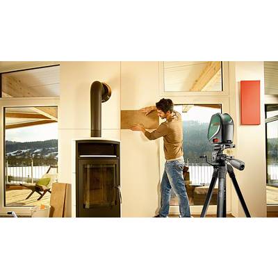 Ein Mann hängt ein Holzstück an eine Wand neben einem modernen Kaminofen. Im Vordergrund steht ein Laser-Werkzeug auf einem Stativ.