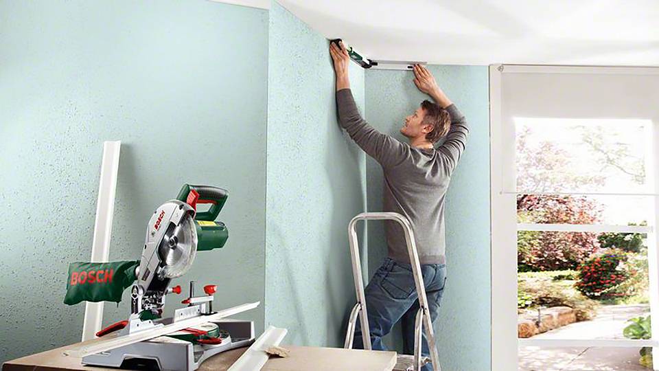 Ein Mann steht auf einer Leiter und misst eine Wand ab. Neben ihm steht eine Gehrungssäge. Im Hintergrund ist ein Fenster mit Gartenblick.