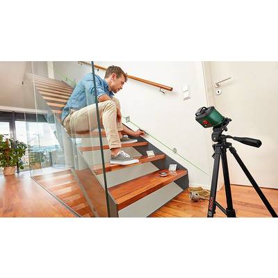 Ein Mann misst mit einem Laser auf einer Holztreppe. Ein Stativ mit Gerät steht daneben. Moderne, helle Umgebung.