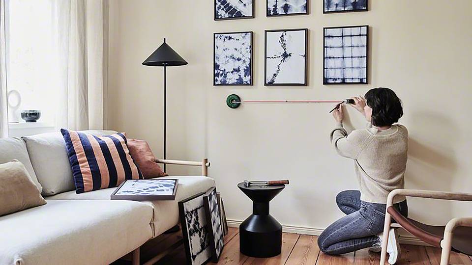 Person misst eine Wohnzimmerwand, um Bilderrahmen aufzuhängen. Die Szene zeigt ein modern eingerichtetes Wohnzimmer mit Sofa und Dekoration.