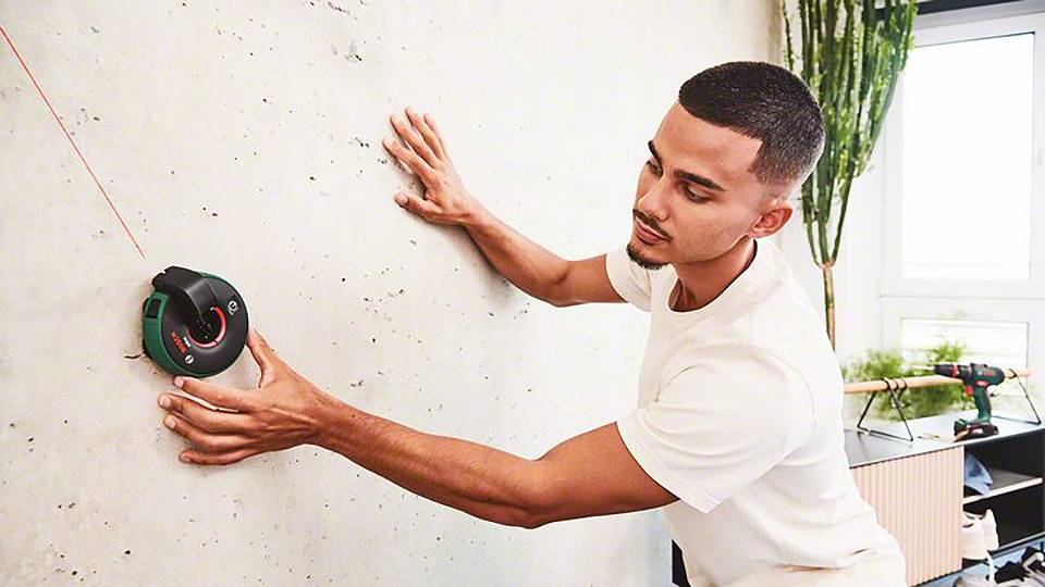 Ein Mann verwendet ein Laserwerkzeug, um eine Linie an der Wand auszurichten. Im Hintergrund sind Pflanzen und ein Fenster zu sehen.