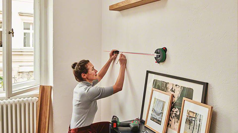 Eine Person misst mit einem Laser-Messgerät eine Wand für die Bildaufhängung in einem Wohnzimmer aus. An der Wand lehnen Bilder.