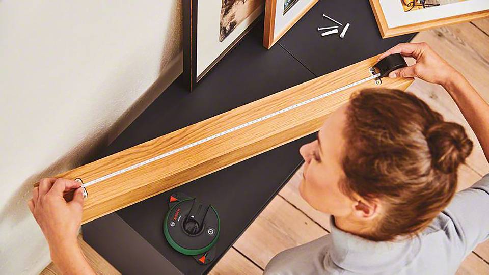 Eine Person befestigt ein Holzbalken an der Wand und nutzt ein digitales Werkzeug zur Ausrichtung. Auf dem Balken liegt ein Zollstock.