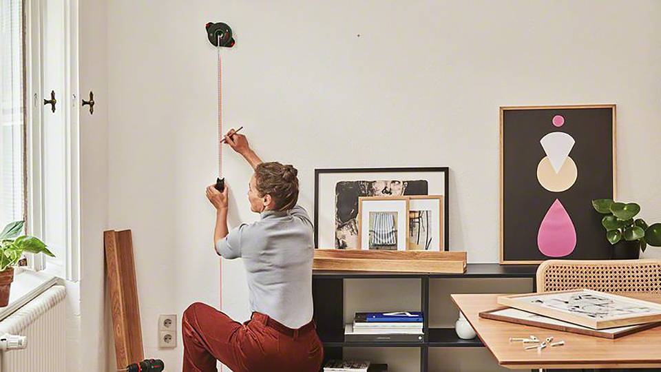 Eine Person misst mit einem Maßband an einer weißen Wand in einem modern eingerichteten Raum, an dessen Wand Bilder und Pflanzen dekorativ platziert sind.