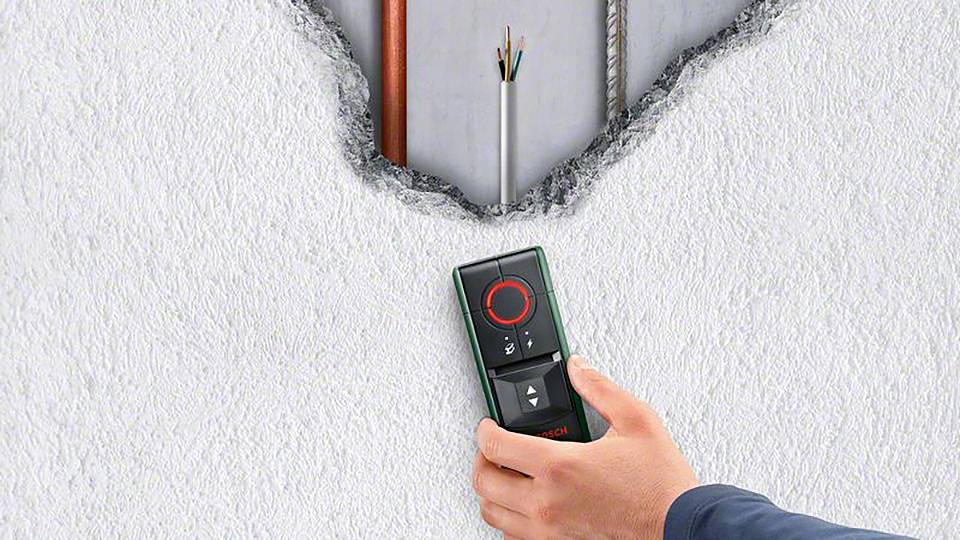Eine Person hält ein Bosch-Messgerät an eine Wand mit sichtbaren Kabeln und Rohren im Inneren, die durch die aufgebrochene Oberfläche sichtbar sind.