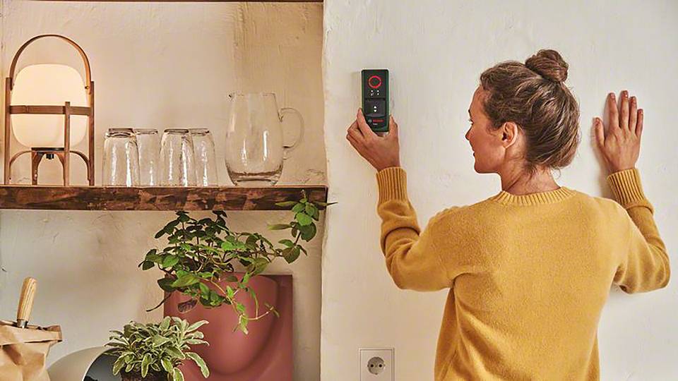 Eine Frau benutzt ein digitales Messgerät, um eine Wand zu vermessen. Im Hintergrund sind eine Lampe und einige Pflanzen auf einem Regal.
