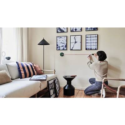 Person misst Wand für Bilderrahmen in einem Wohnzimmer aus. Aufgeräumter Raum mit Sofa, Lampen und Holzboden.