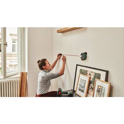 Eine Frau misst mit einem Lasermessgerät an einer weißen Wand die Position für Regale. Auf dem Boden liegen eingerahmte Bilder und Werkzeug.