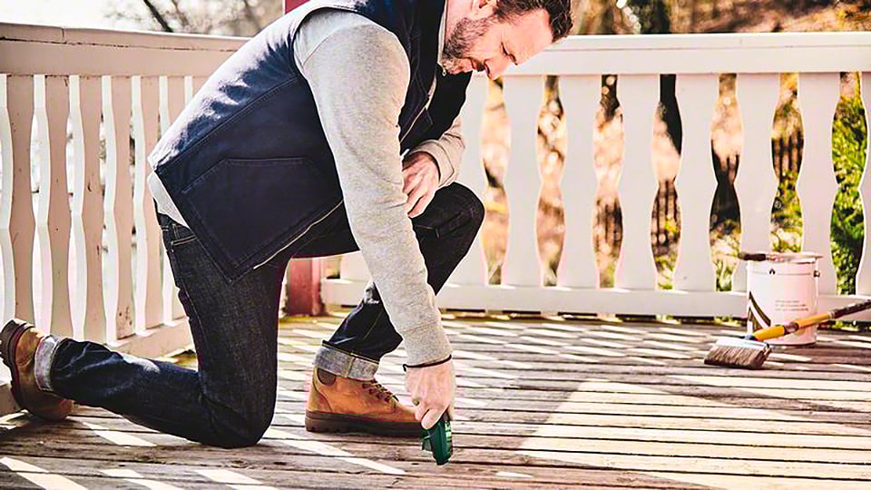 Ein Mann kniet auf einer Holzveranda und verwendet ein Werkzeug, um den Boden zu bearbeiten. Im Hintergrund steht ein Farbeimer.