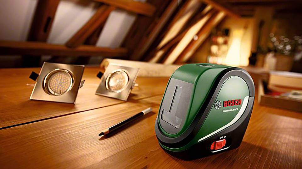 Ein grüner Bosch-Laser-Entfernungsmesser liegt auf einem Holztisch, daneben ein Bleistift und zwei quadratische LED-Leuchten.