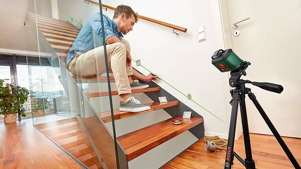Ein Mann verwendet ein Laser-Werkzeug auf einer Holztreppe zur genauen Vermessung. Im Hintergrund sind Glasgeländer und ein Stativ zu sehen.