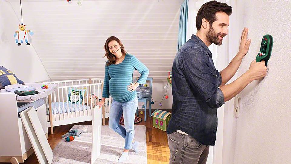 Ein Paar richtet ein Kinderzimmer ein: Eine schwangere Frau steht lächelnd, während ein Mann ein Gerät an der Wand benutzt.