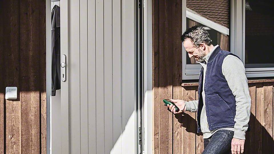 Ein Mann steht vor einer Holztür und benutzt ein Smartphone. Die Sonne scheint und er trägt eine Weste über einem grauen Pullover.