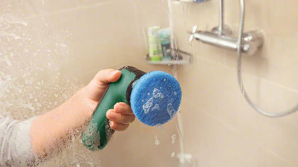 Eine Hand hält ein Reinigungsgerät mit einem blauen Schwamm unter einer Dusche. Wasser läuft über die Fliesen.